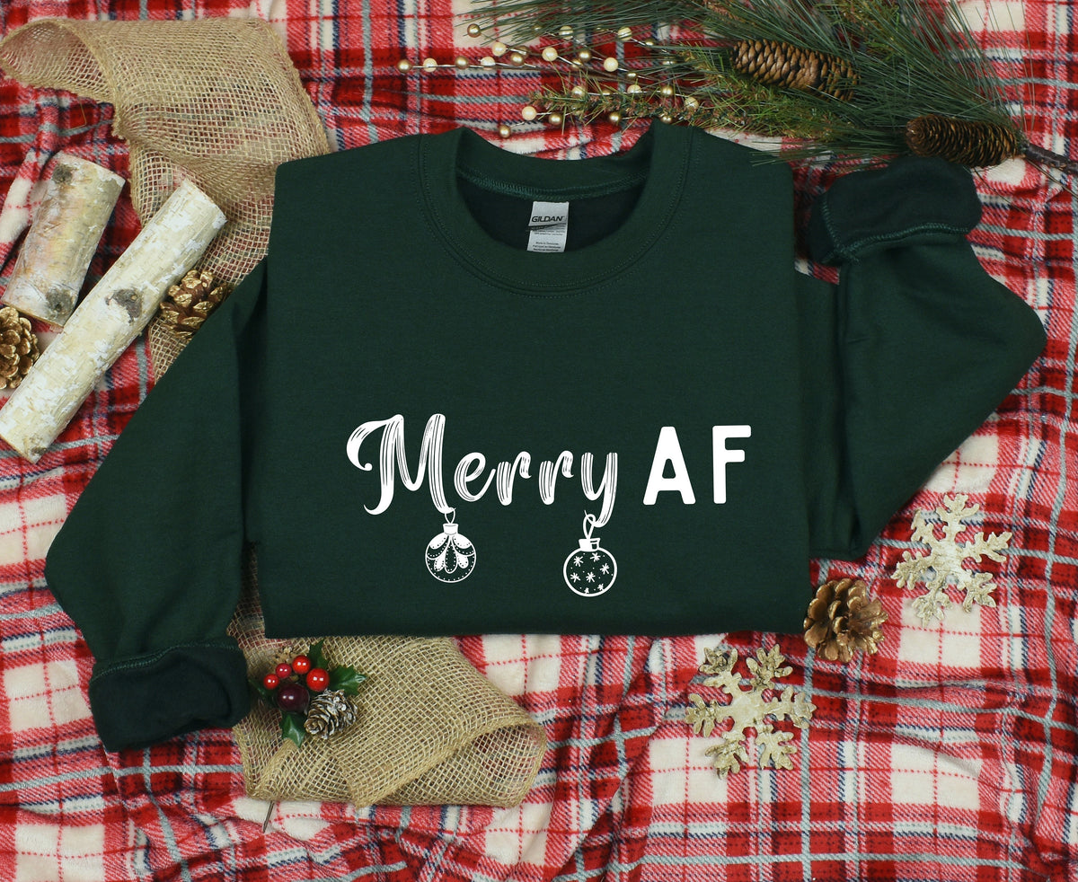 Merry AF Christmas Sweatshirt in forest green laid flat on red plaid with pinecones, birch, burlap and greenery.