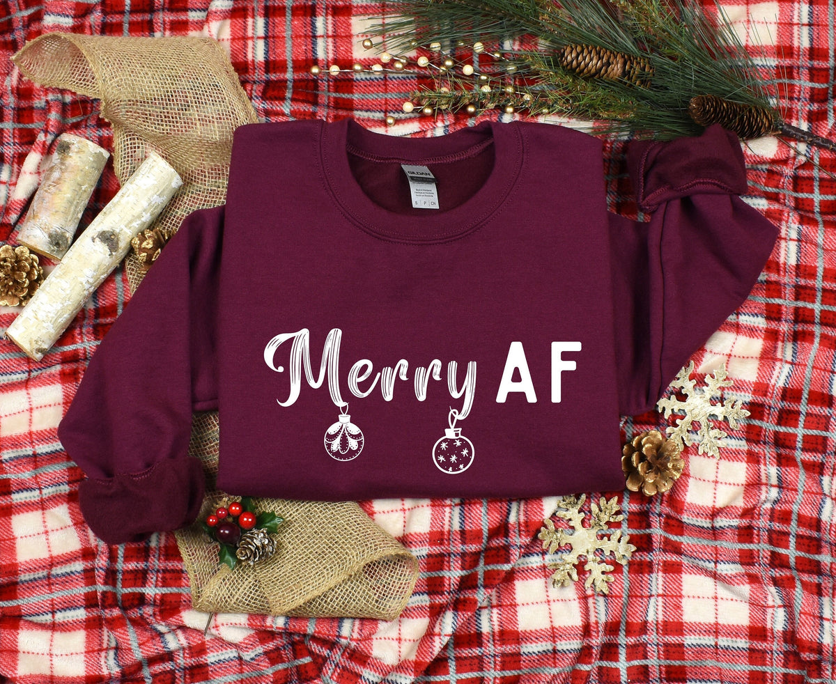 Merry AF Christmas Sweatshirt in maroon, shown flat on red plaid with pine greenery, cones, birch logs, burlap and snowflakes.