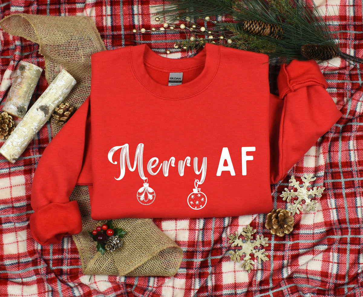 Merry AF Christmas Sweatshirt in red, overhead view on plaid with pine branches, pinecones, burlap and snowflakes.