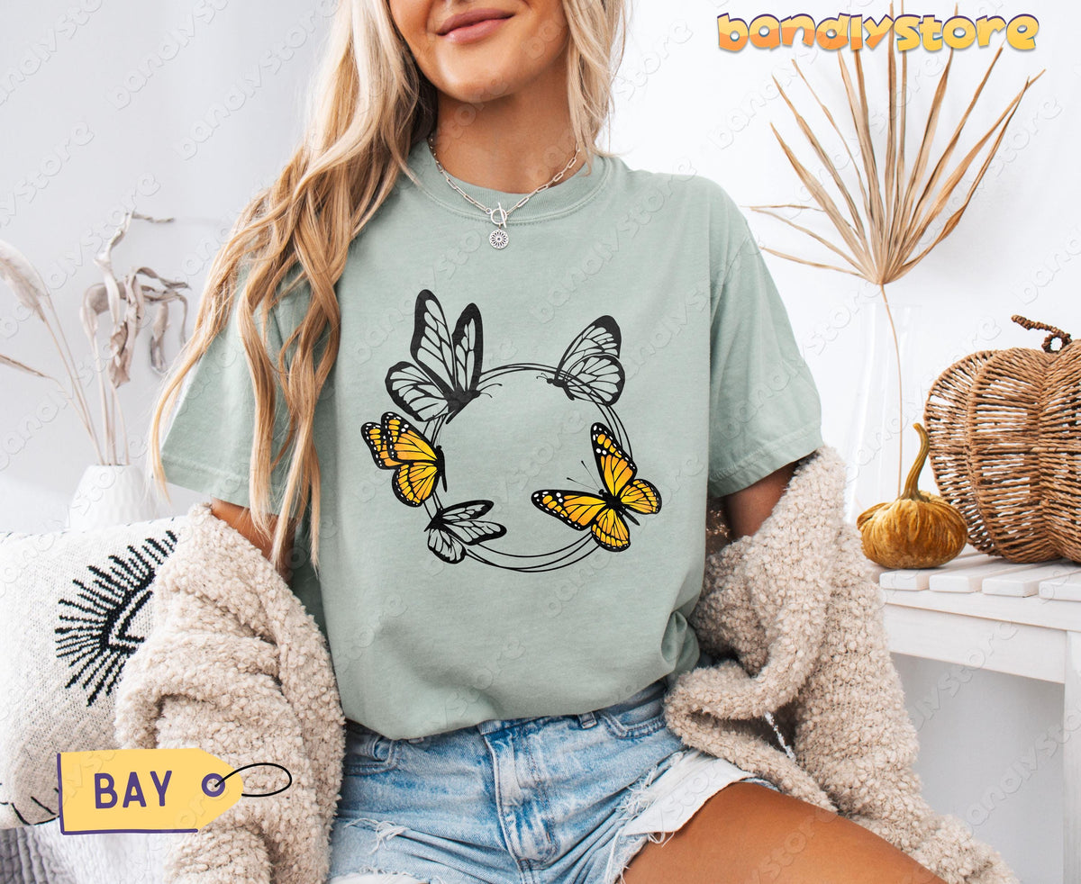 Monarch Butterfly Wreath T-Shirt in Bay on a seated model, front view beside dried plants, pumpkin and basket.