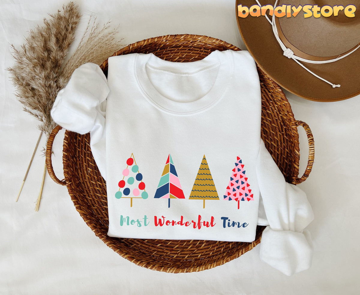 Most Wonderful Time Christmas Trees Sweatshirt white, folded in a round wicker basket beside dried grass and a brown hat.