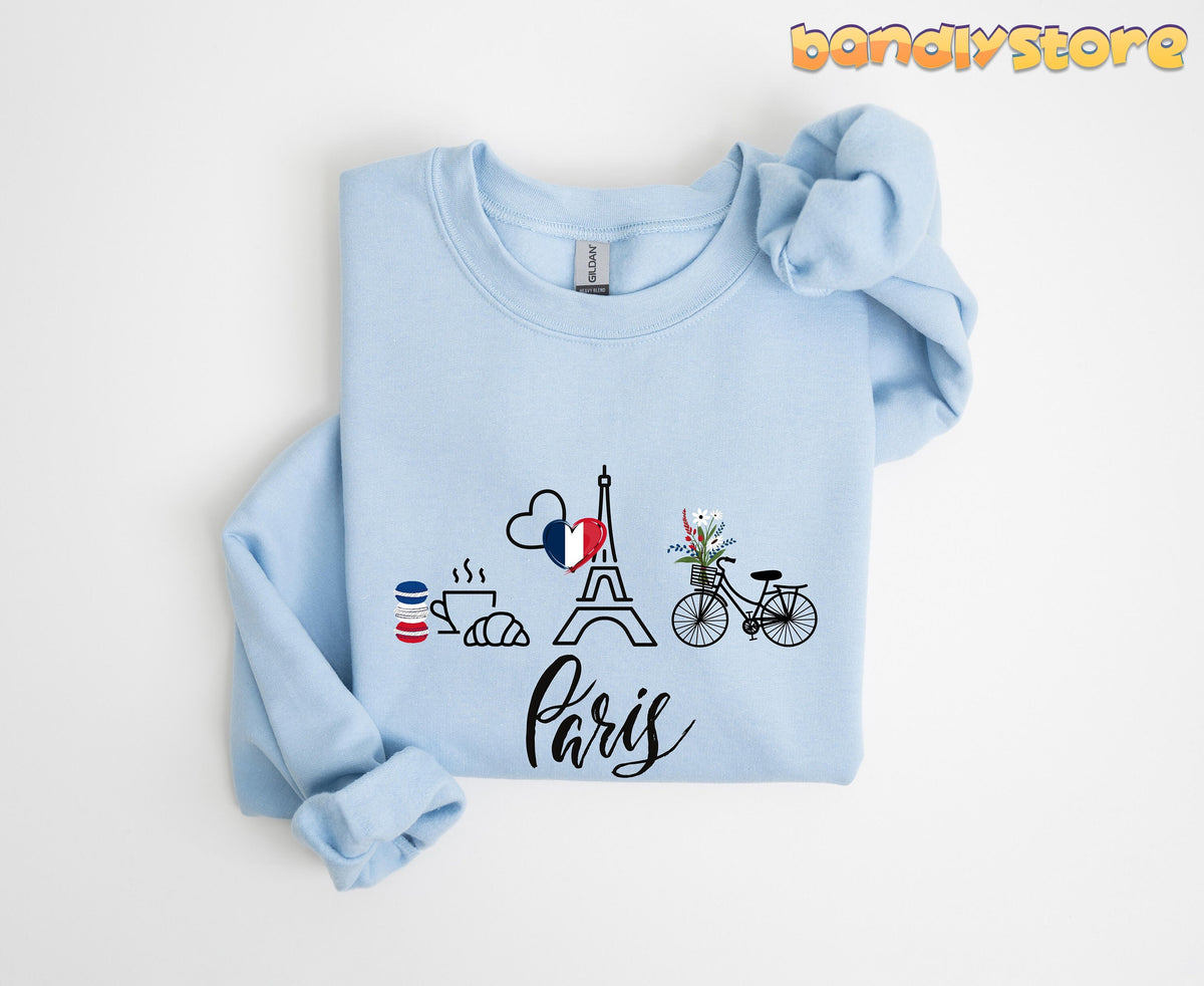 Paris Eiffel Tower Sweatshirt light blue folded flat with macarons coffee croissant Eiffel Tower bike flowers Paris script.