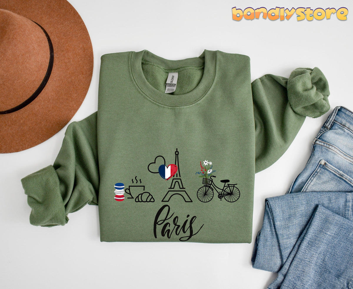 Paris Eiffel Tower Sweatshirt in military green, folded flat beside a brown hat and blue jeans, showing Paris icons.