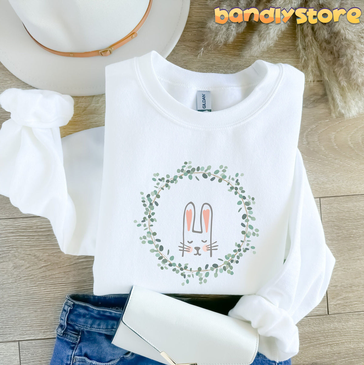 Spring Wreath Bunny Sweatshirt in white, flat-lay on wood with hat, jeans, clutch and dried grass.