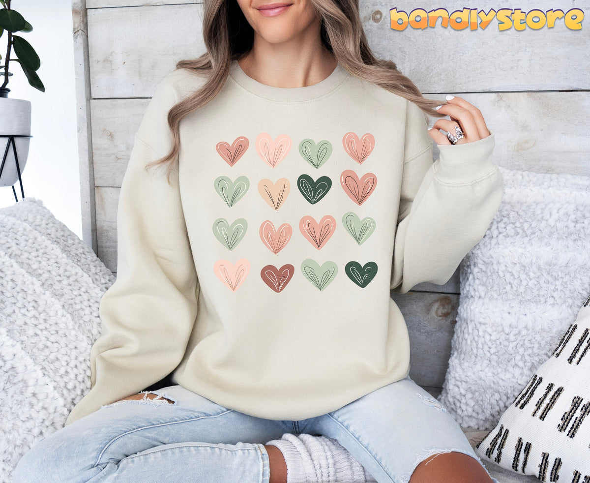 Cute Pastel Hearts Sweatshirt in sand, worn seated against wood wall with pillows and plant nearby.