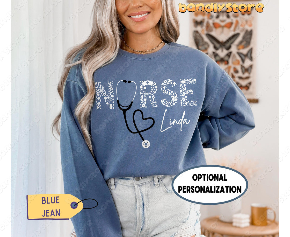Personalized Nurse Sweatshirt in Blue Jean worn front-facing by a woman in a room with a butterfly poster and table.
