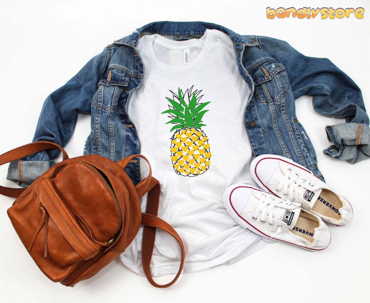Pineapple T-Shirt in white laid flat over a denim jacket next to a brown backpack and sneakers.