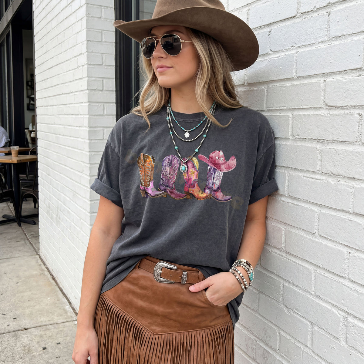 Pink Cowgirl Boots T-Shirt in pepper on a woman leaning against a white brick wall.