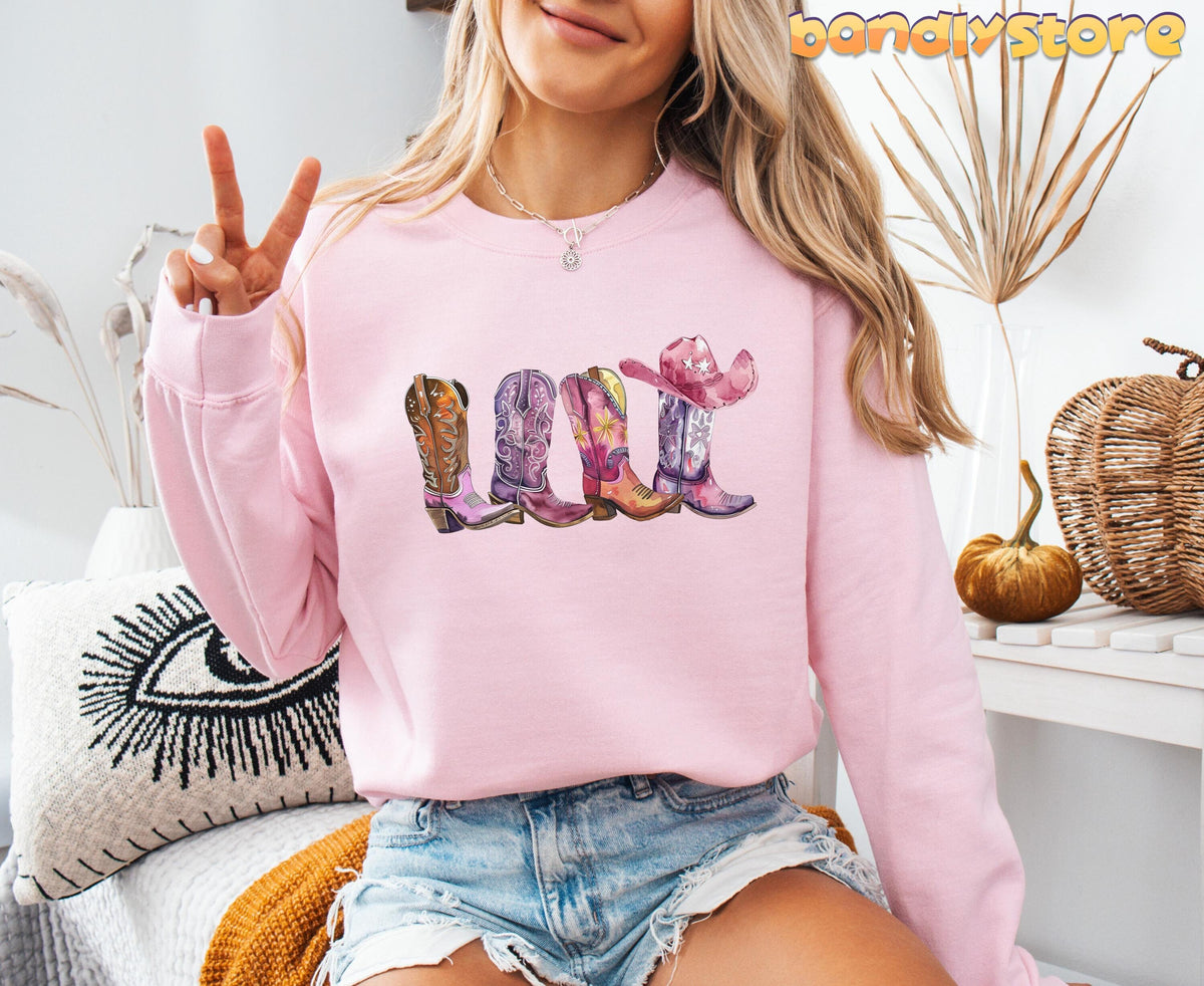 Pink Cowgirl Boots Sweatshirt light pink crewneck shown front-on with four illustrated cowgirl boots, indoor props behind.