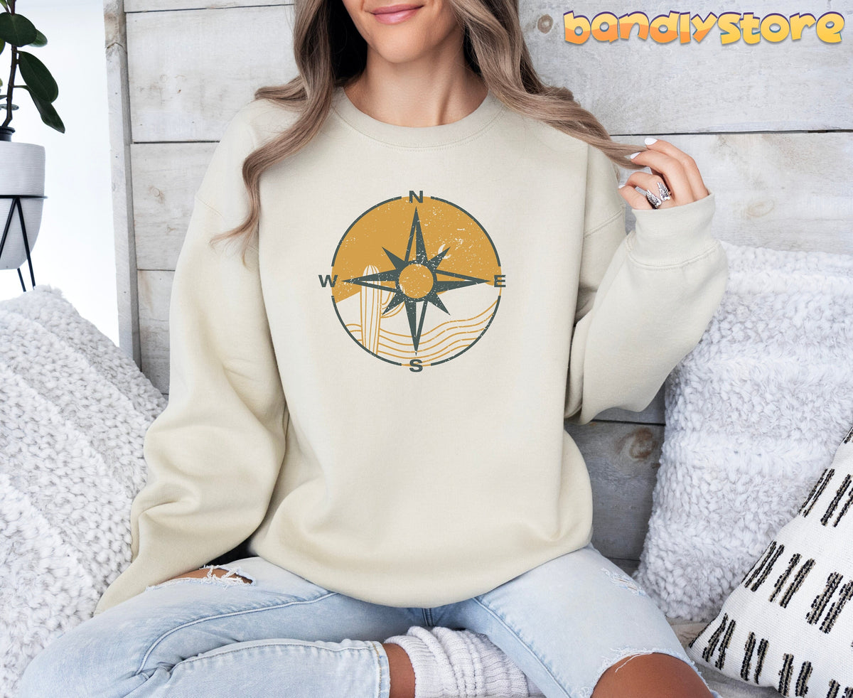 Retro Compass Sweatshirt in sand on a seated woman against a wood wall with white pillows and a plant.