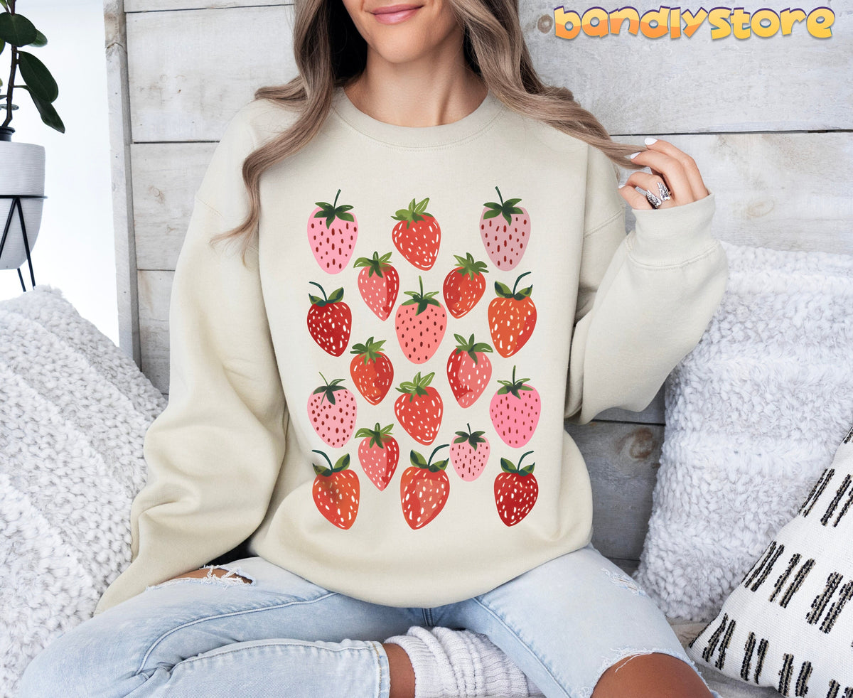 Retro Strawberry Sweatshirt in sand, worn front-facing by a seated model amid pillows and a wooden wall.