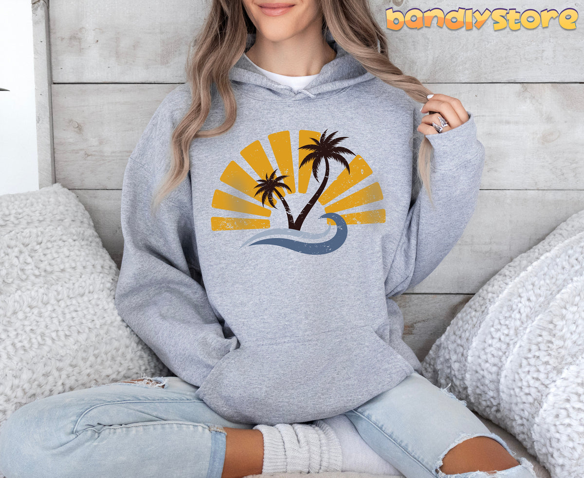 Retro Sunset Hoodie in grey on a seated person facing forward amid white pillows by a wood wall.