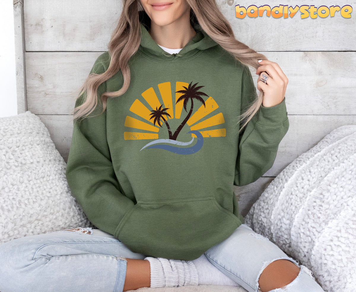 Retro Sunset Hoodie in military green, worn by a seated model among white pillows against a wood wall.
