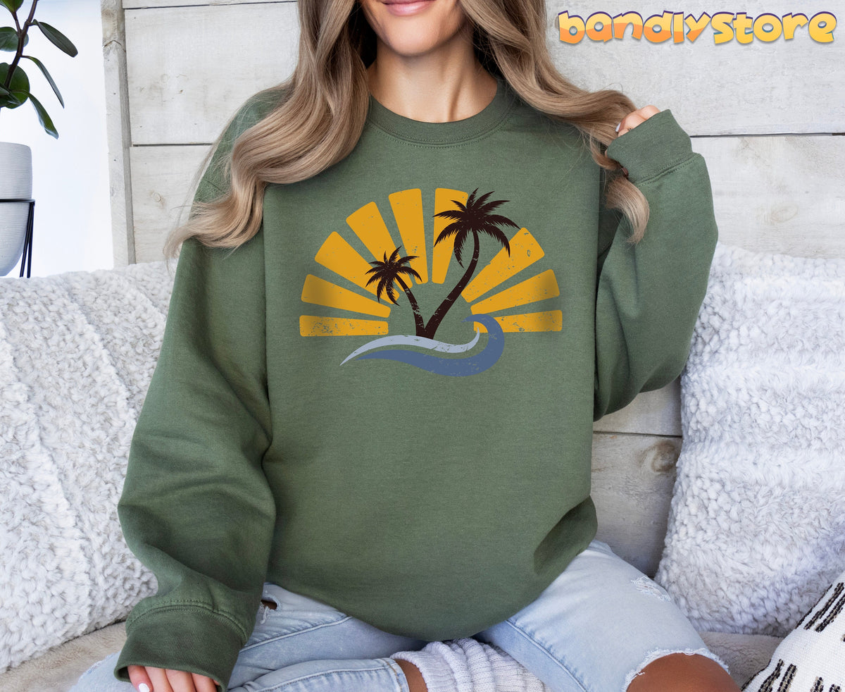 Retro Sunset Sweatshirt in military green on a seated person facing forward amid white pillows and wood wall.