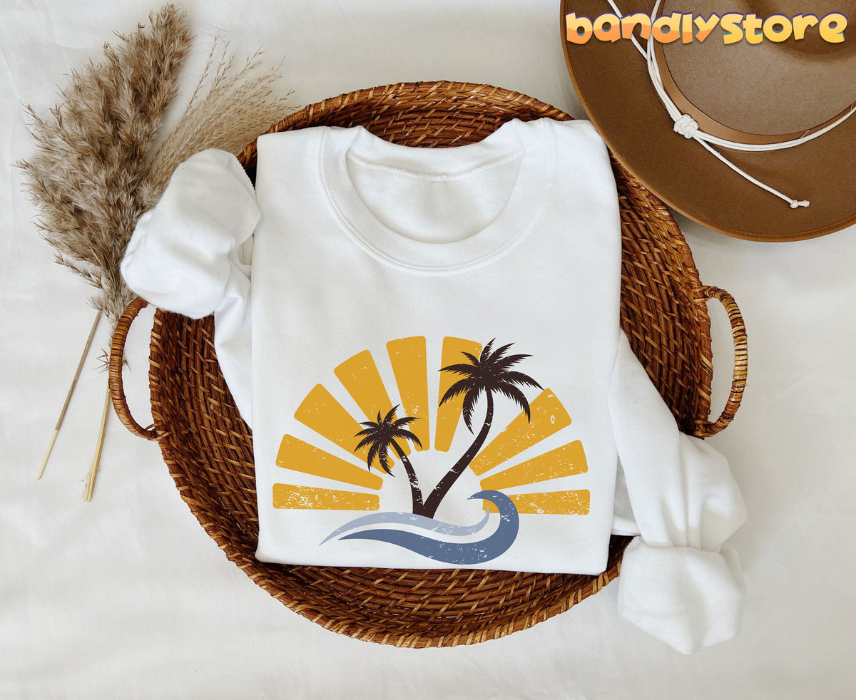 Retro Sunset Sweatshirt in white folded in a woven basket beside pampas grass and a hat, overhead view.