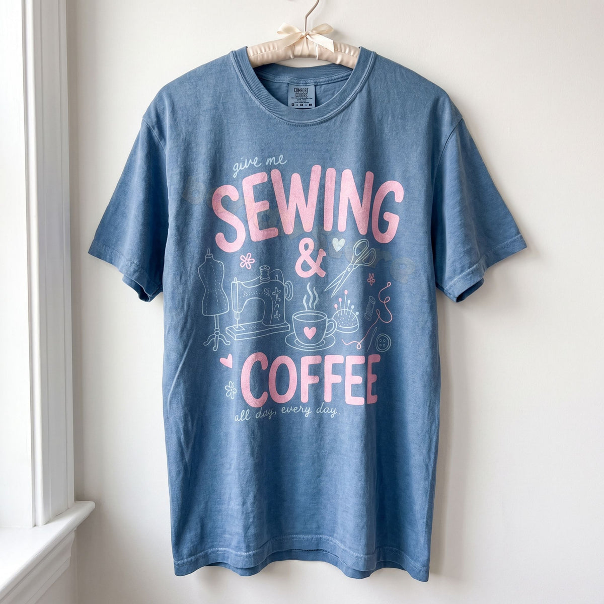 Sewing And Coffee T-Shirt in Blue Jean hanging on a hanger against a white wall by a window.