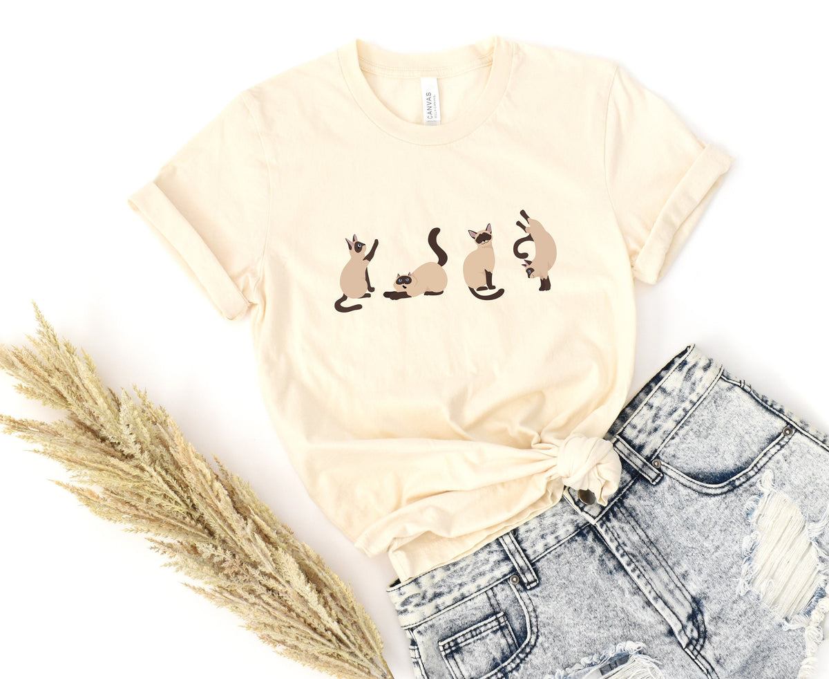 Siamese Cat T-Shirt in Natural, flat-laid with four cat graphics, dried grass left and knotted hem over ripped jeans.