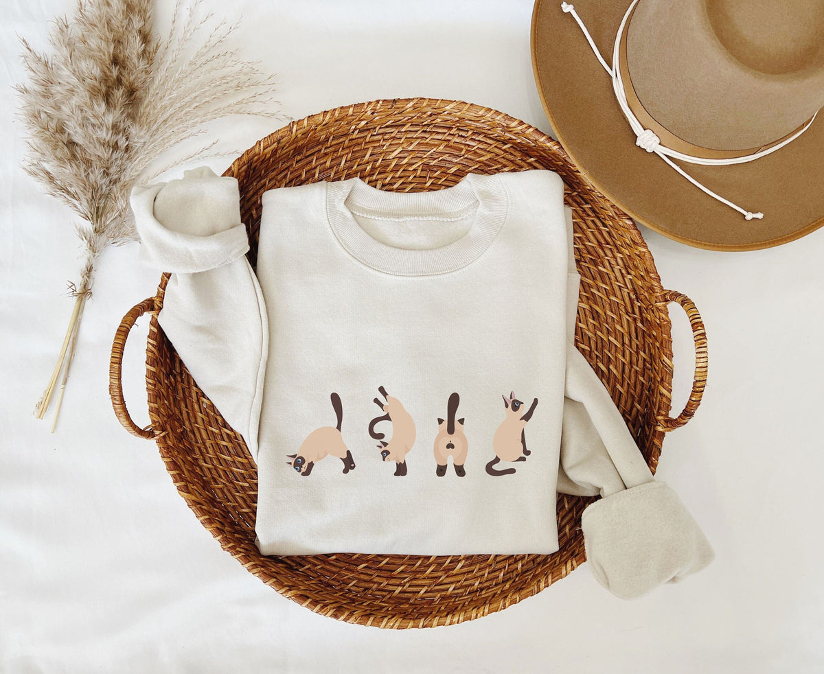 Siamese Cat Butts Sweatshirt in sand folded in a wicker basket beside pampas grass and a hat.