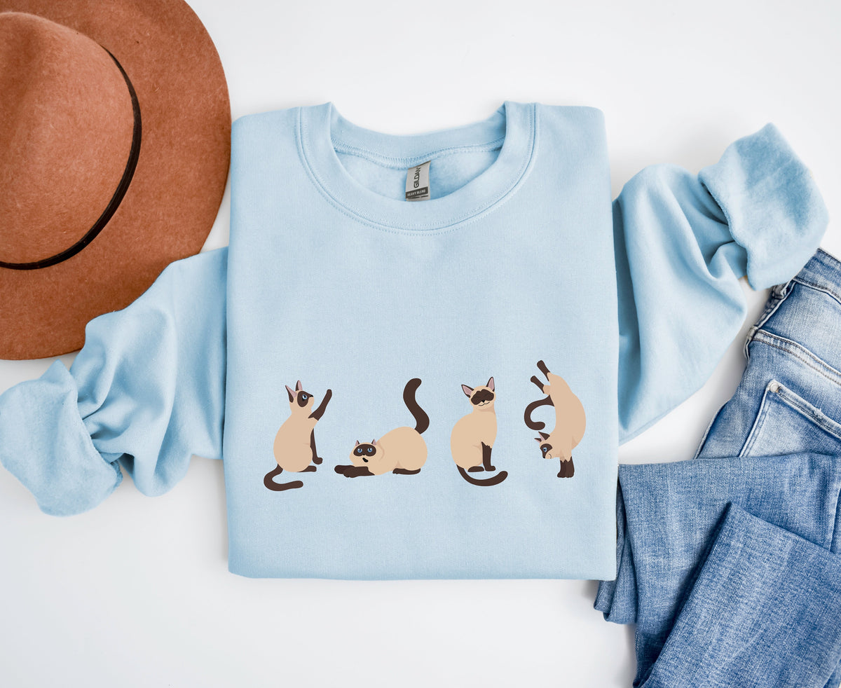 Siamese Cat Sweatshirt in light blue folded flat beside a brown hat and jeans.