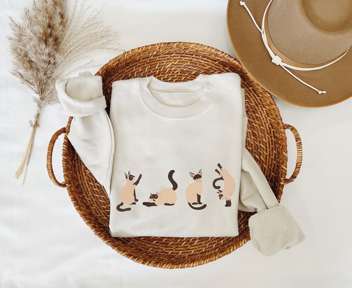Siamese Cat Sweatshirt in sand folded in a woven basket beside pampas grass and a hat, top-down view.