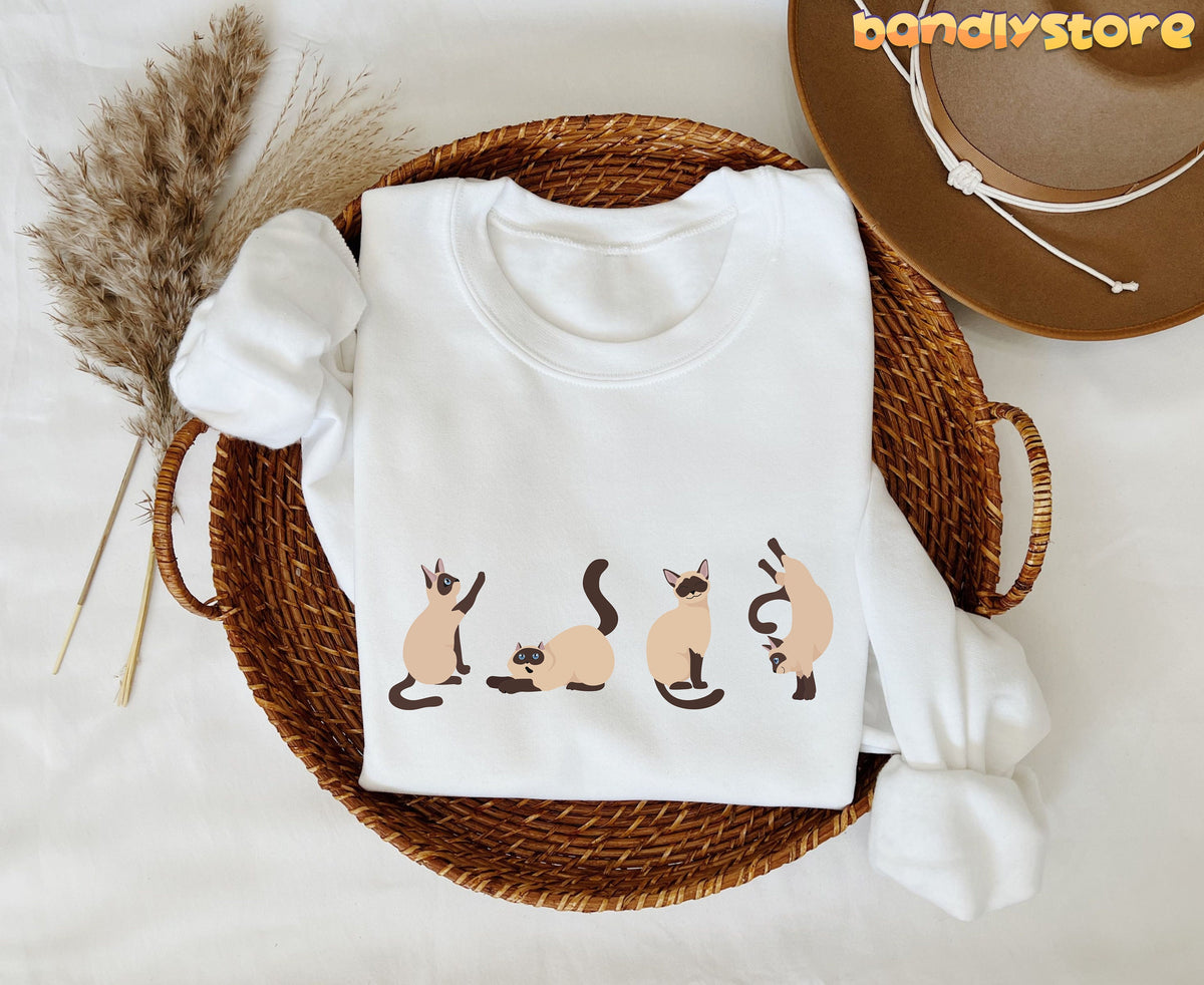 Siamese Cat Sweatshirt white folded in a wicker basket beside pampas grass and a brown hat.