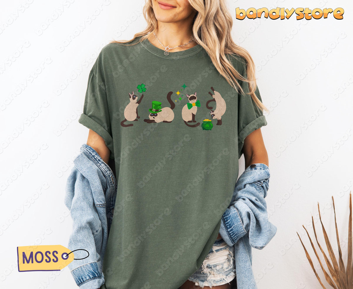 Siamese Cat Shamrock T-Shirt in moss on model against white wall, denim jacket held, plant nearby.