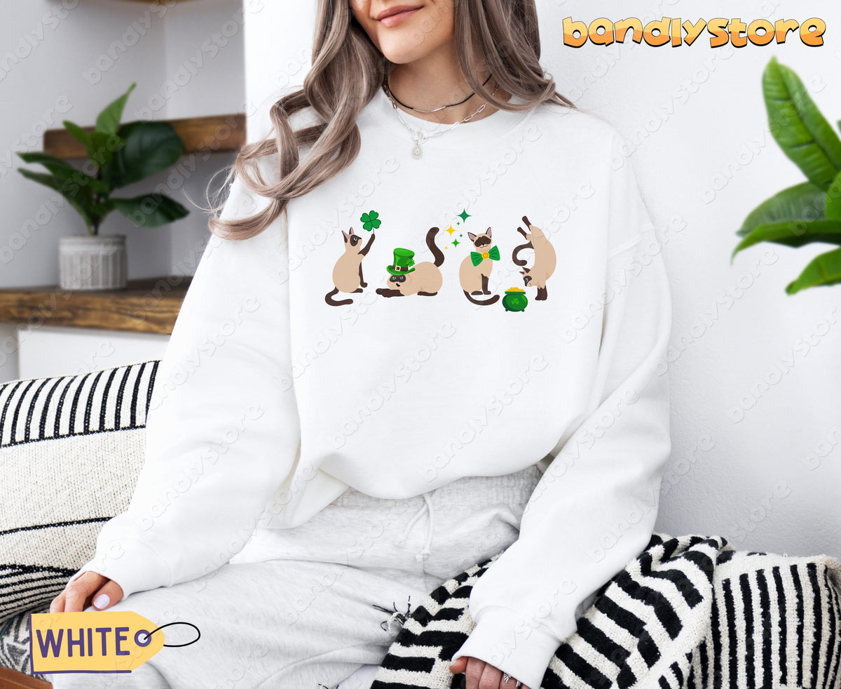 Siamese Cat St Patrick's Day Sweatshirt in white, worn seated indoors amid shelves, plants and striped cushions.