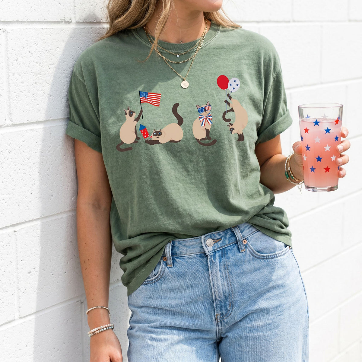 Siamese Cats 4th of July T-Shirt in moss worn by a person leaning against a white brick wall holding a glass.