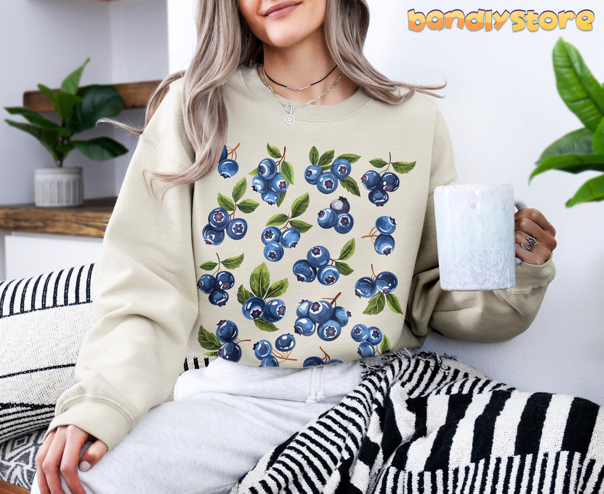 Blueberry Sweatshirt in sand on seated person holding mug amid plants and striped blanket.