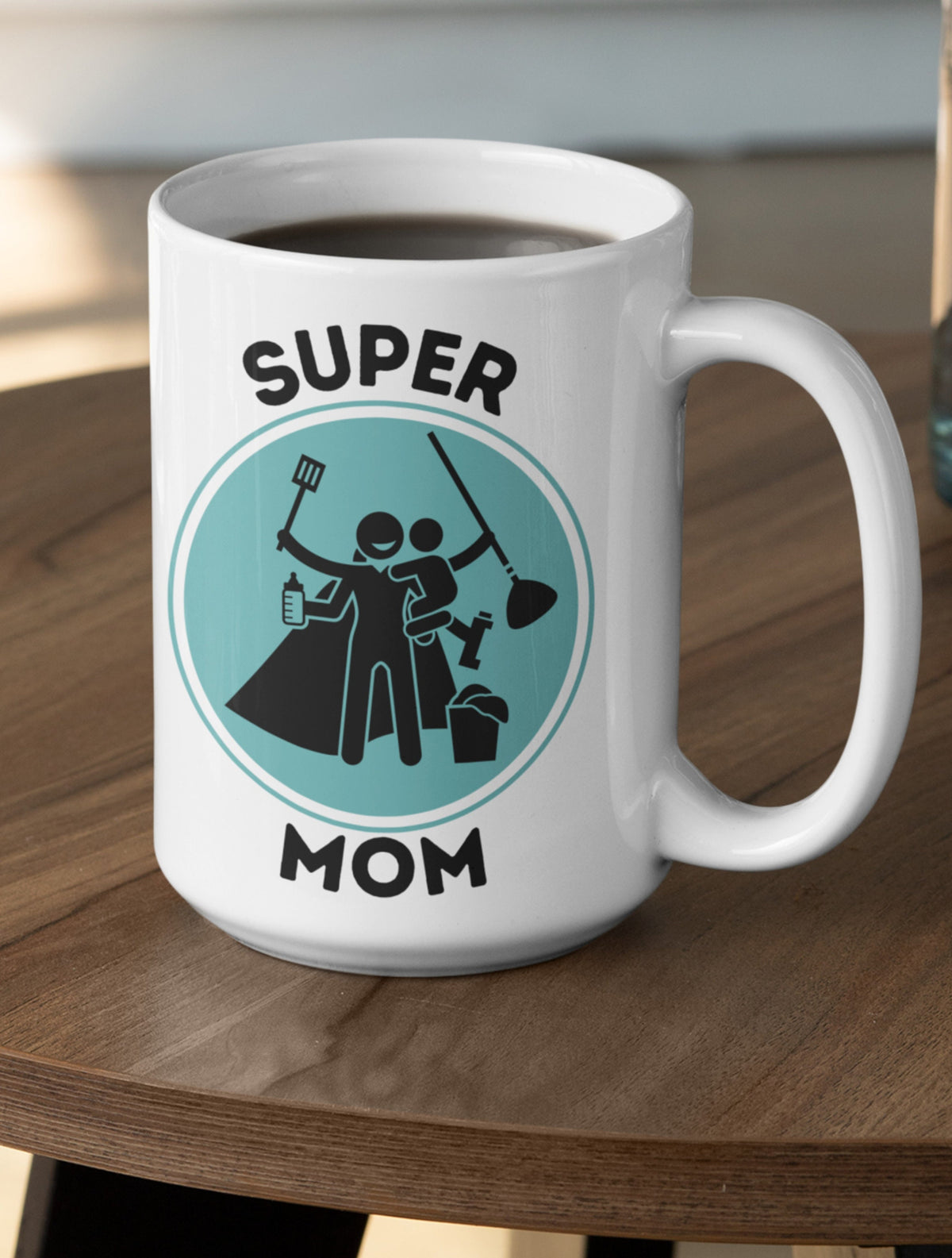 Supermom Mug on a wood table, filled with dark liquid, teal circle graphic of mom with baby and tools.