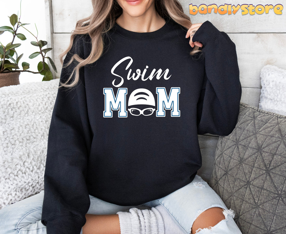 Swim Mom Sweatshirt in black worn front-facing by a seated woman beside a plant and pillows.