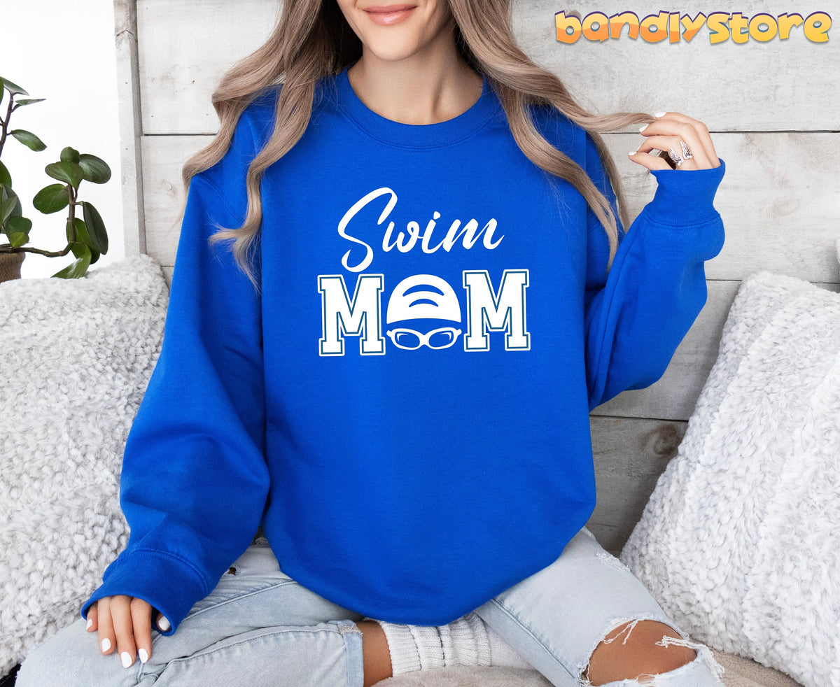 Swim Mom Sweatshirt in royal on a woman sitting on a couch with white pillows and a plant, front view.