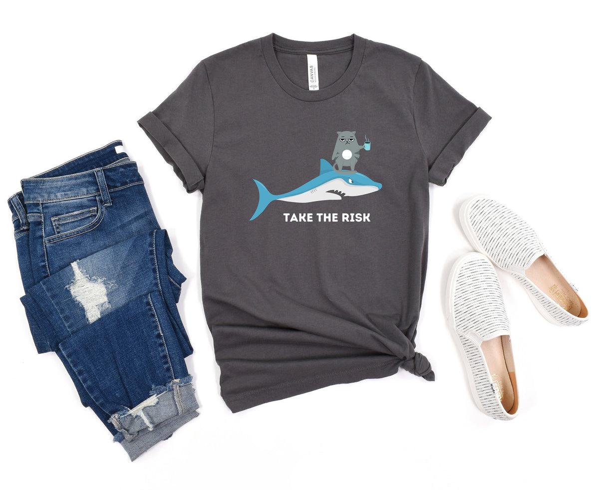 Take The Risk Funny Cat T-Shirt in dark grey, flat-laid with ripped blue jeans left and white slip-ons right.