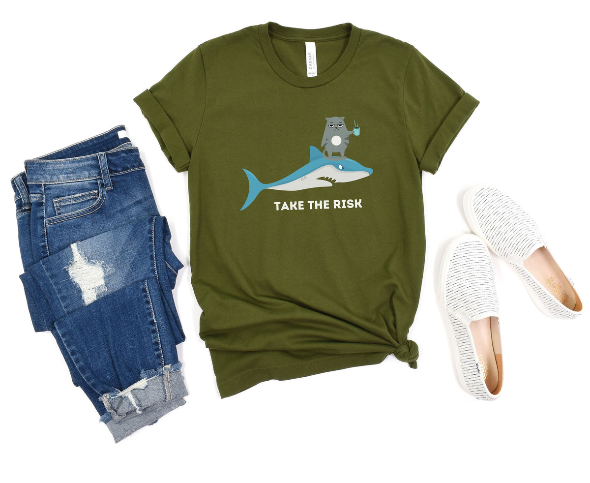Take The Risk Funny Cat T-Shirt in olive laid flat beside blue ripped jeans and white slip-on shoes.