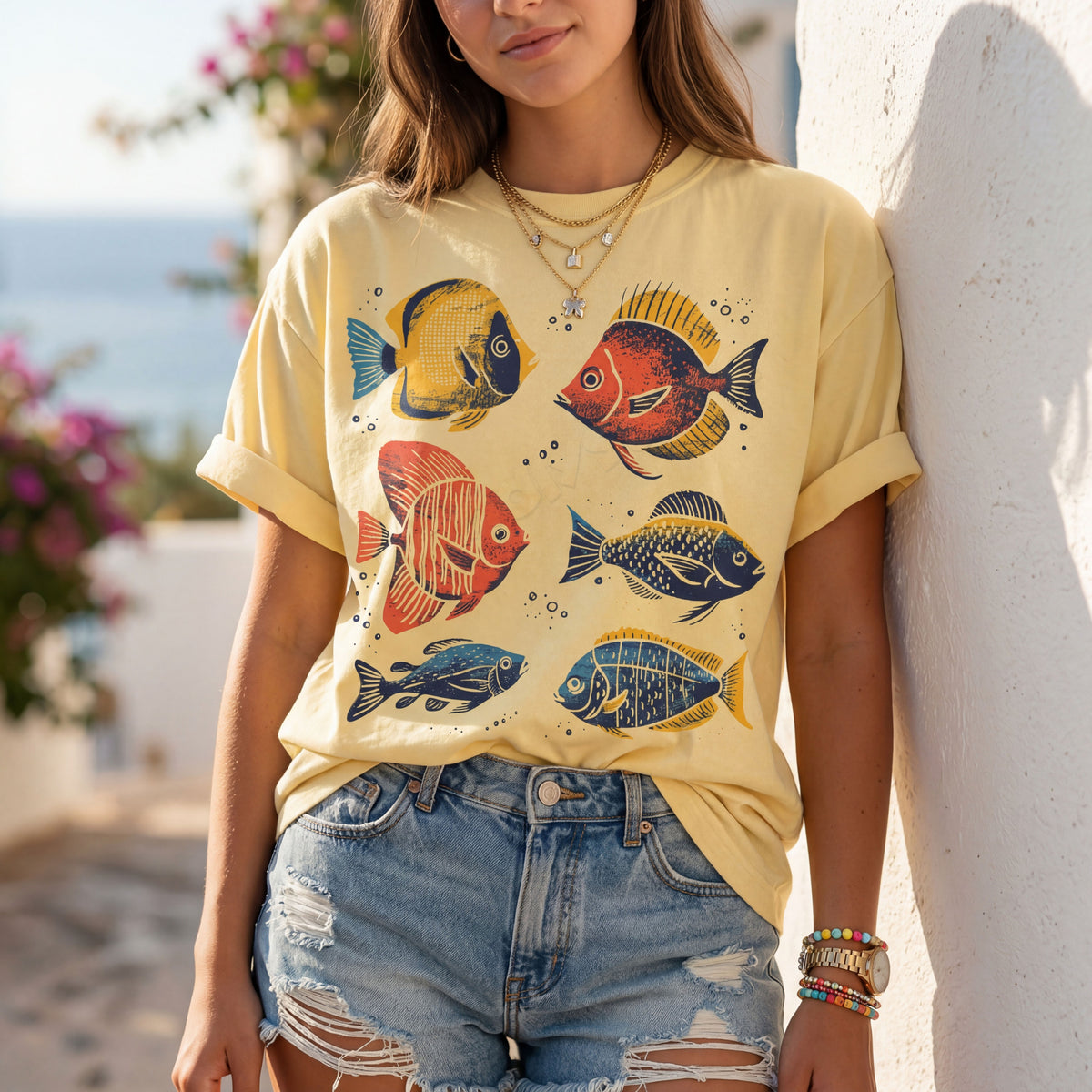 Tropical Fish T-Shirt in Butter, worn front-on outdoors by a woman against a white wall with sea and flowers behind.