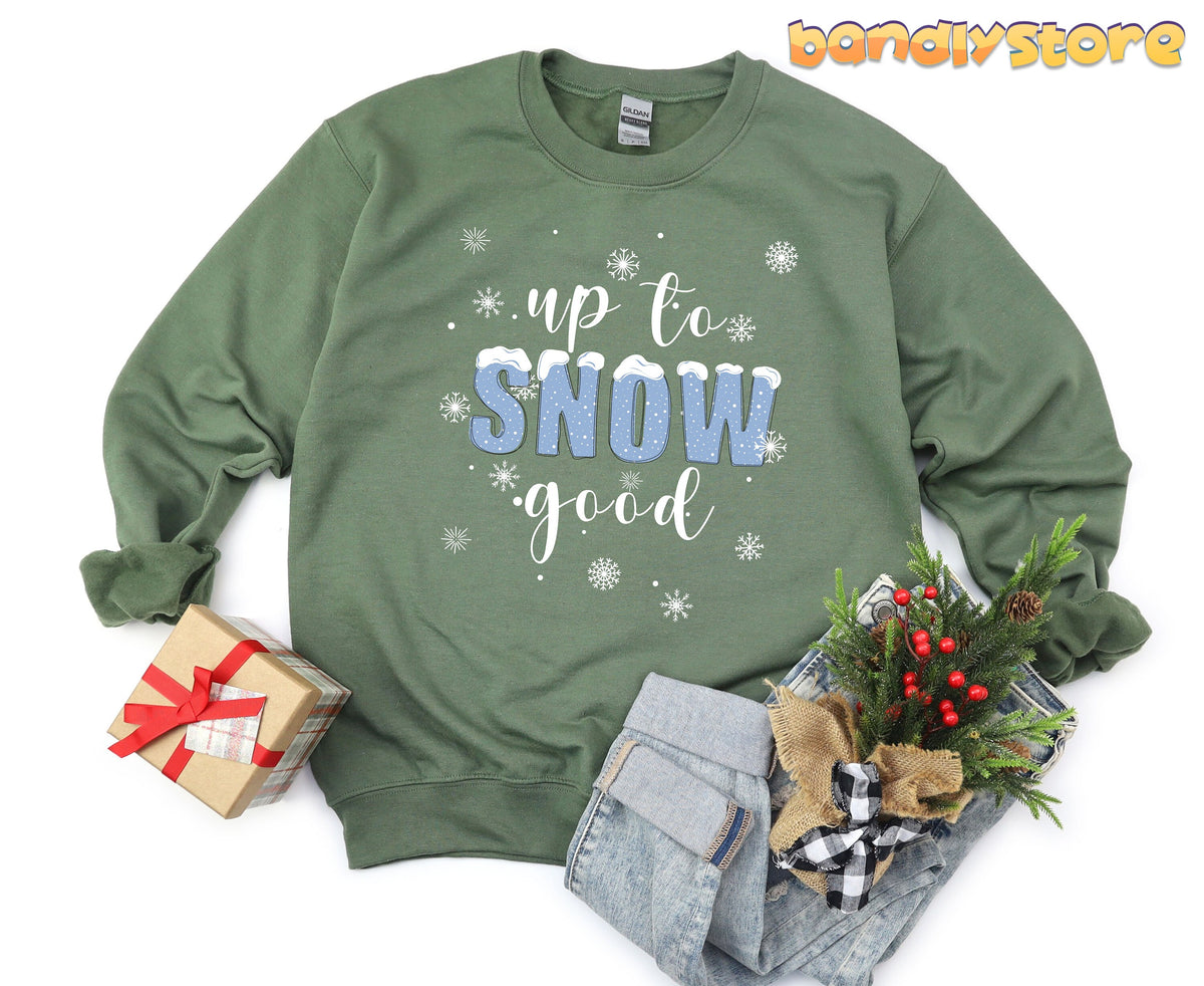 Up To Snow Good Sweatshirt in military green laid flat beside a gift box and jeans with greenery.
