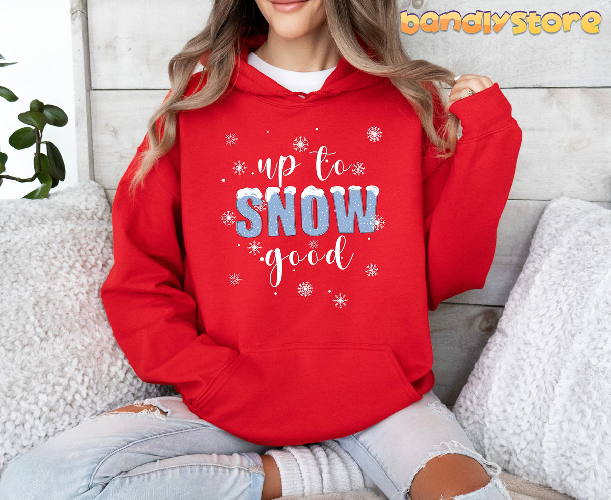 Up To Snow Good Hoodie in red, worn by a seated person amid white pillows, a plant, and wood wall.