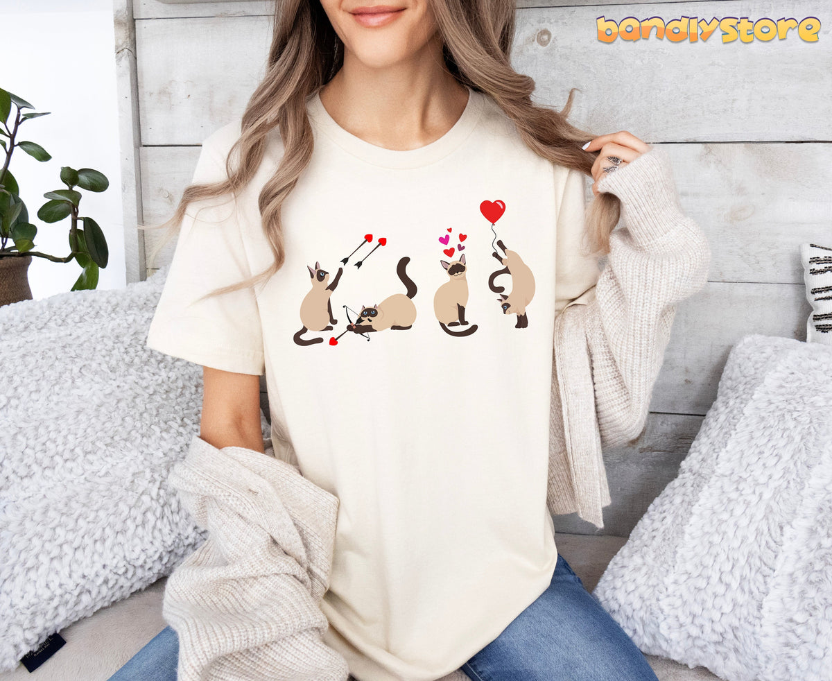 Valentine's Day Siamese Cat T-Shirt in natural front-facing on seated person amid pillows, plant and wooden wall.