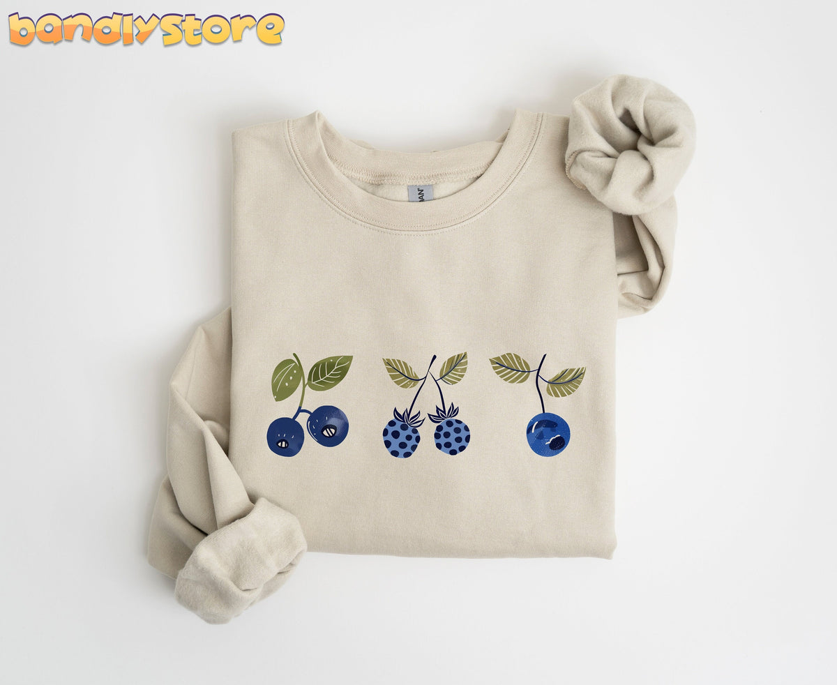 Vintage Blue Berries Sweatshirt in sand, folded flat on white with sleeves crossed and blueberry print across the chest.