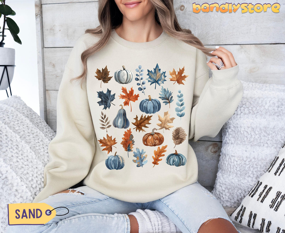 Vintage Fall Leaves Sweatshirt in sand on a seated model amid pillows and a plant by a wooden wall.