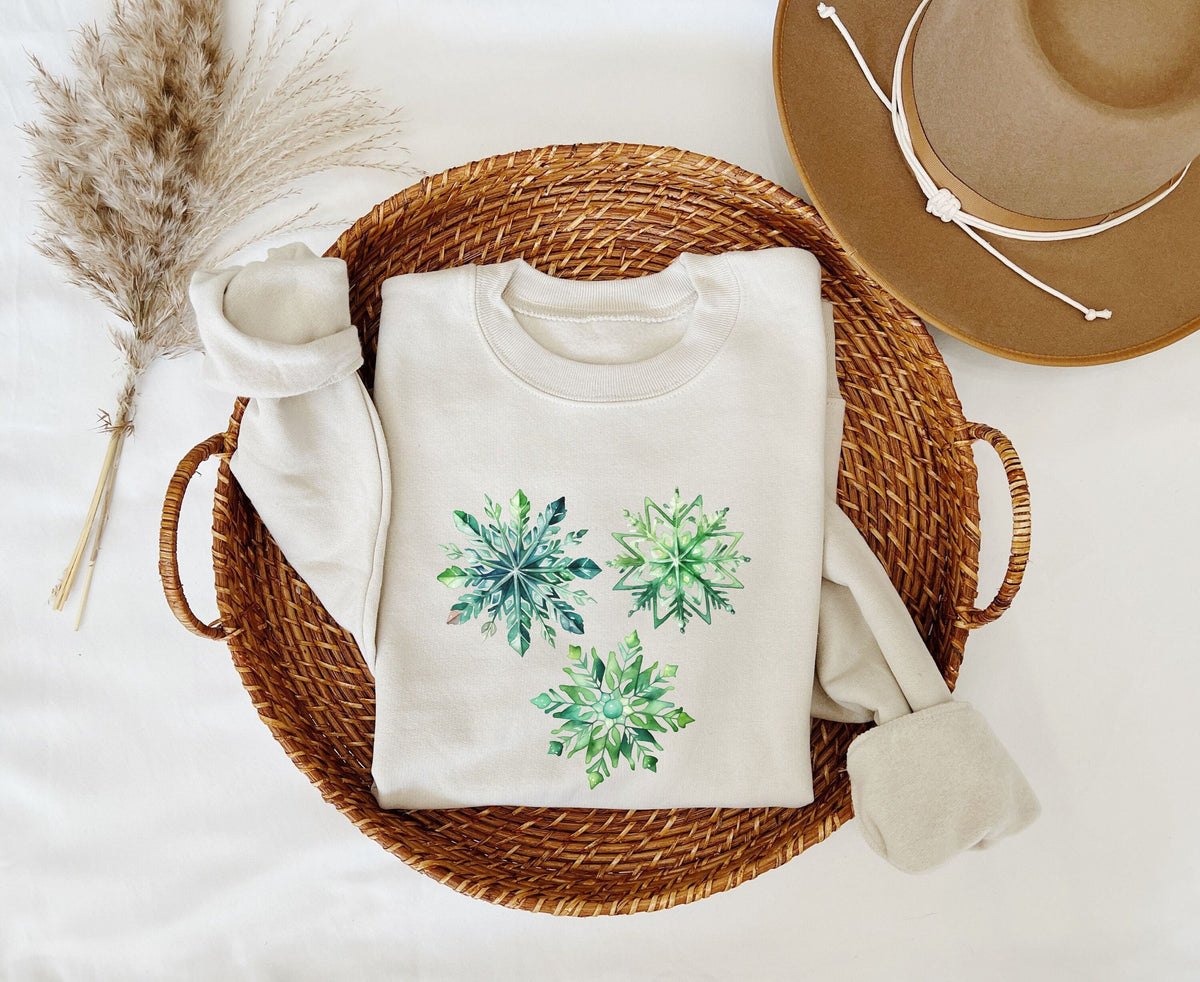 Watercolor Snowflake Sweatshirt in sand folded in a wicker basket with pampas grass and a hat nearby, top-down view.