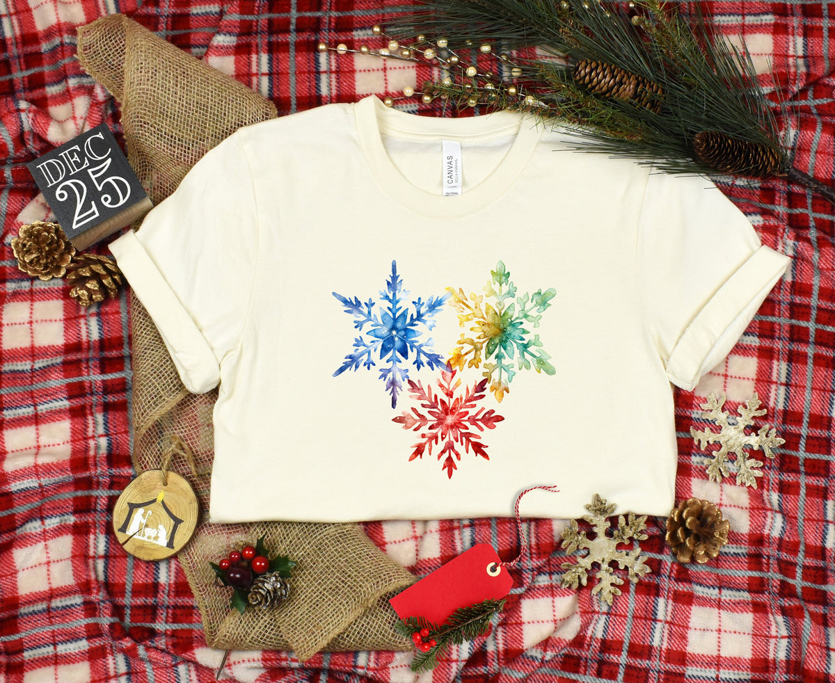 Colorful Watercolor Snowflakes T-Shirt in natural, flat lay on red plaid with pinecones and ornaments.