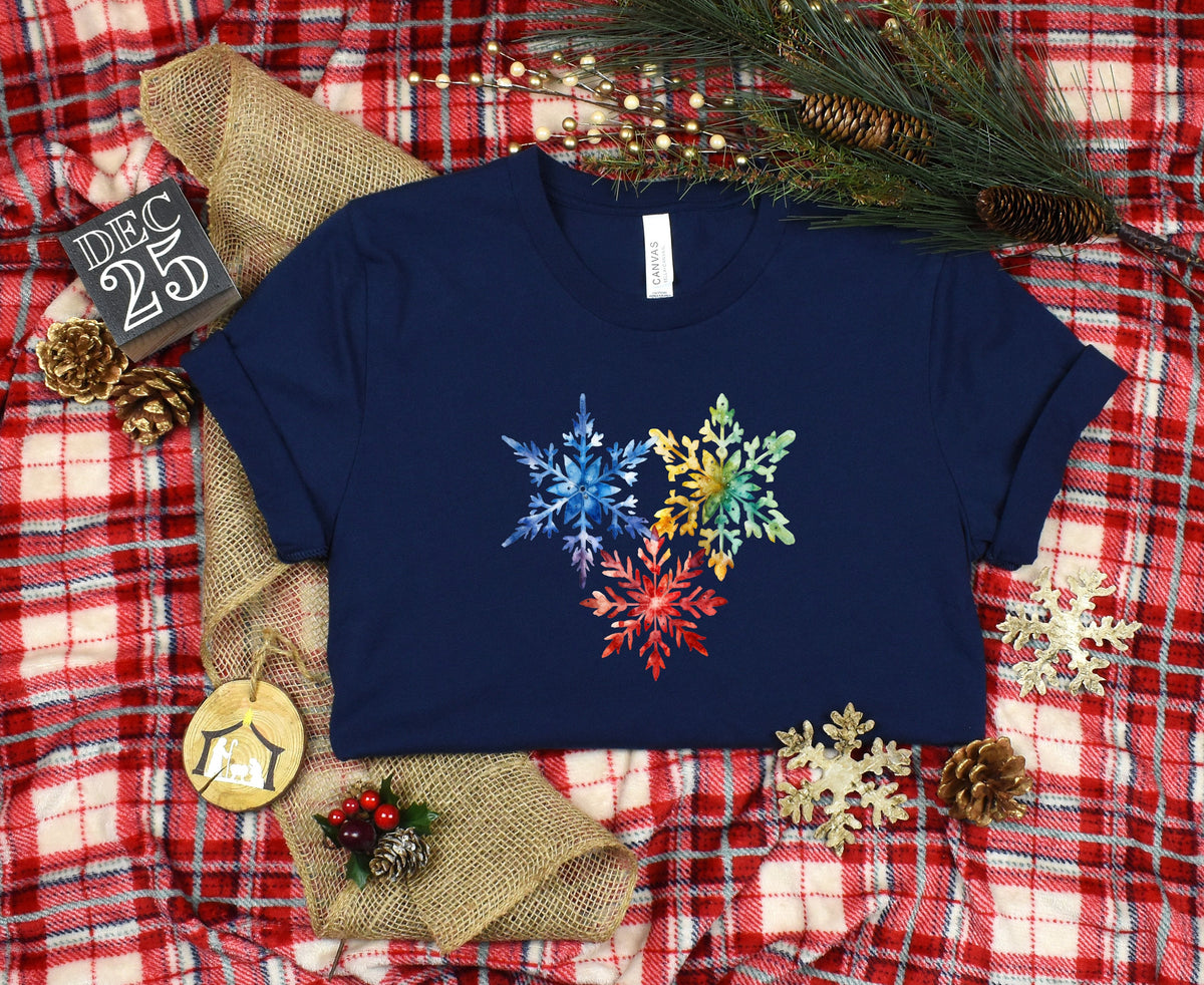 Colorful Watercolor Snowflakes T-Shirt in navy laid flat on red plaid among pinecones, greenery and ornaments.