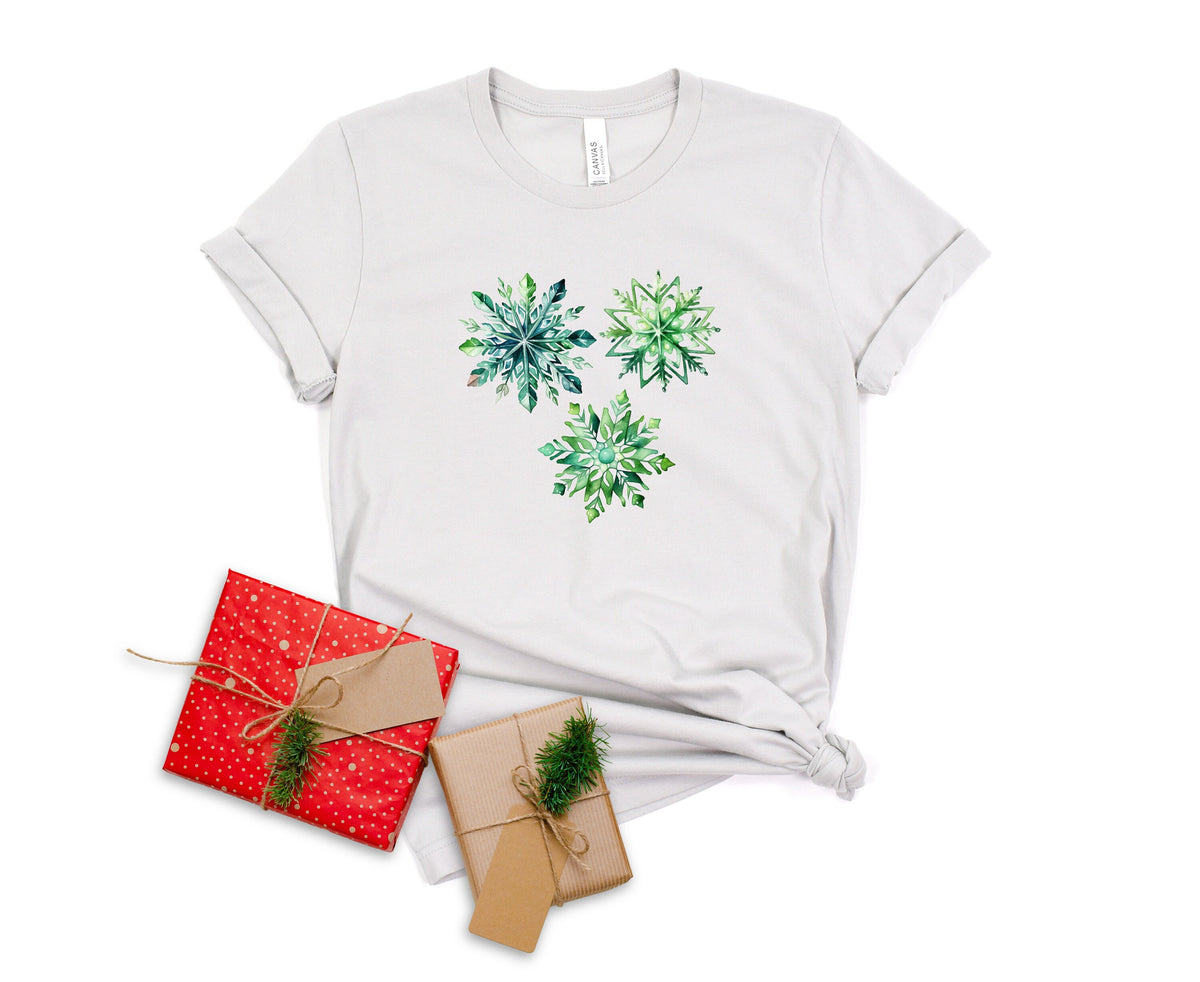 Watercolor Snowflakes T-Shirt in silver laid flat beside two wrapped gifts with greenery.