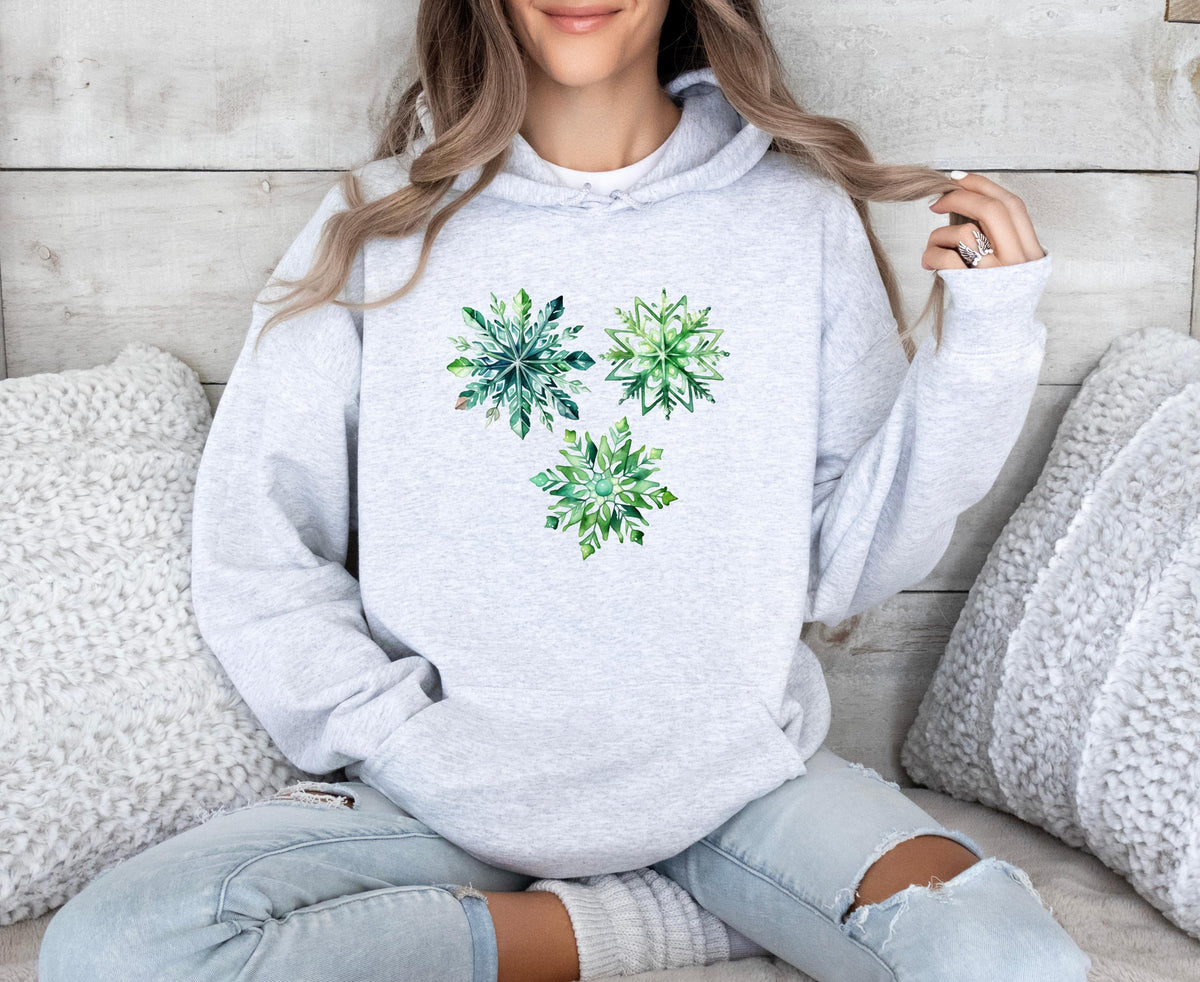 Watercolor Snowflakes Hoodie in ash on a seated person amid fluffy pillows against a wooden wall.