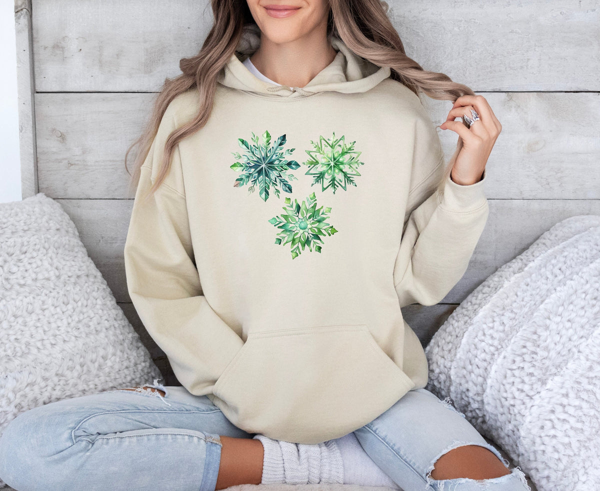 Watercolor Snowflakes Hoodie in Sand on a cross-legged person against a wood wall with white pillows.
