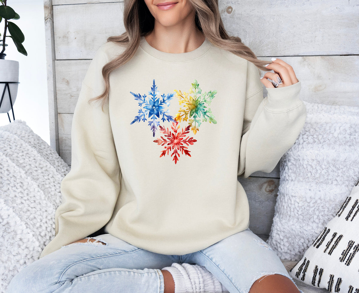 Colorful Watercolor Snowflakes Sweatshirt in sand, worn seated amid pillows against a wood wall.