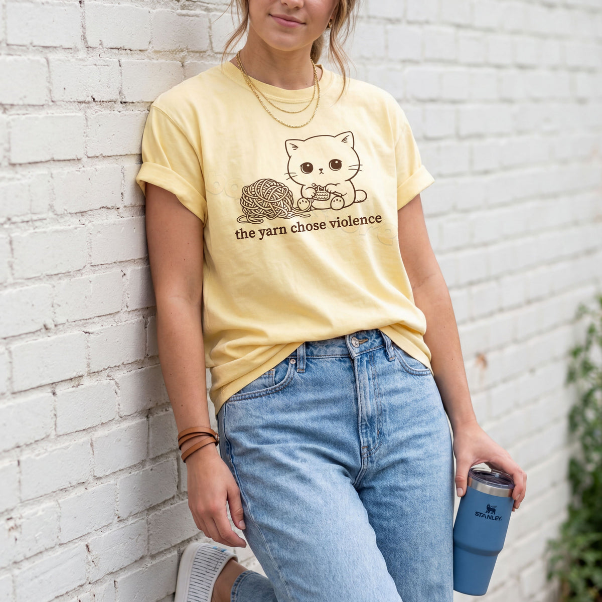 The Yarn Chose Violence T-Shirt in butter on a person leaning against a white brick wall holding a blue tumbler.