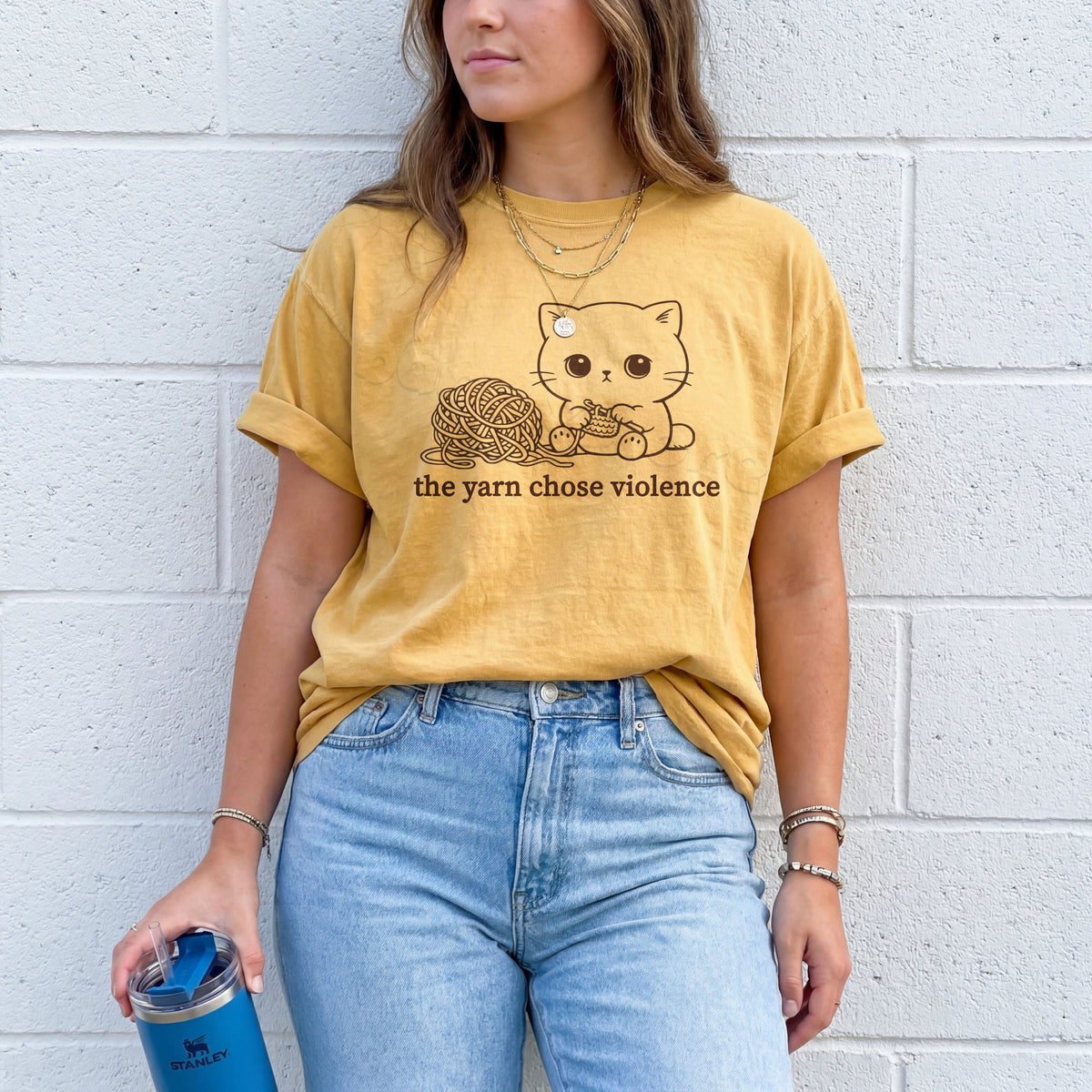 The Yarn Chose Violence T-Shirt in mustard, worn front-facing by a model against a white brick wall with a blue tumbler.