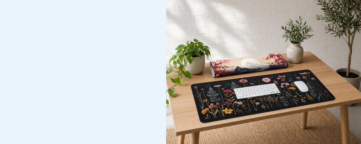 2 printed neoprene desk mats on a tidy desk against a sunlit white brick wall, one in use under a keyboard, one lying flat beside it and one loosely rolled on the desk, with an olive tree